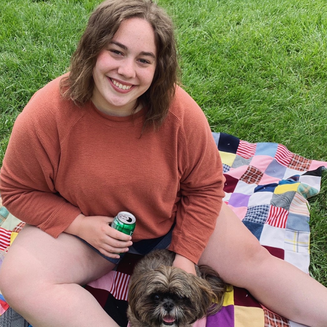 a photo of a female student in a dark orange sweater sitting on a picnic blanket with her small grey dog