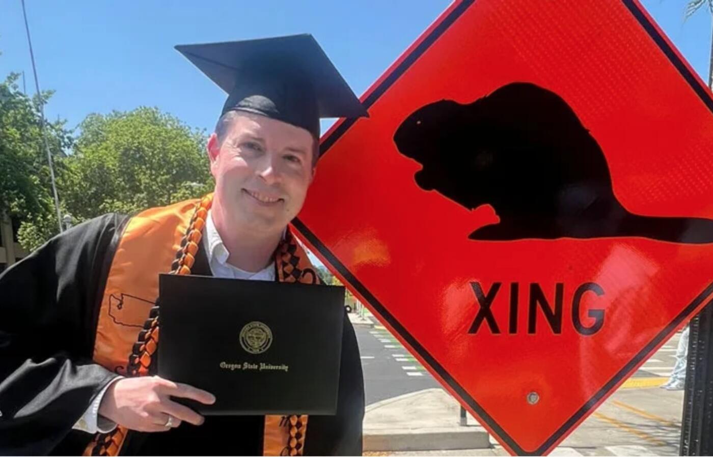 man in graduation cap and gown poses next to beaver crossing sign