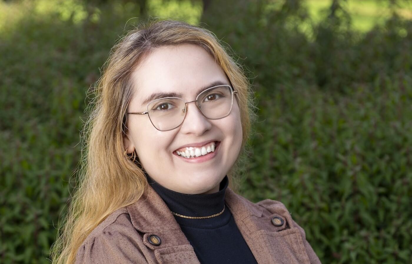 woman with glasses smiling at camera in front of a green bush