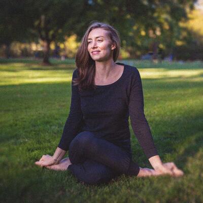 Photo of Kate Gallagher sitting in gomukhasana, a yoga pose, in the grass. She is wearing black pants and a black shirt. 
