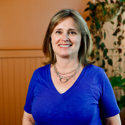 Photo of Angie smiling in front of the plants at Marigold. She is wearing a bright blue shirt.