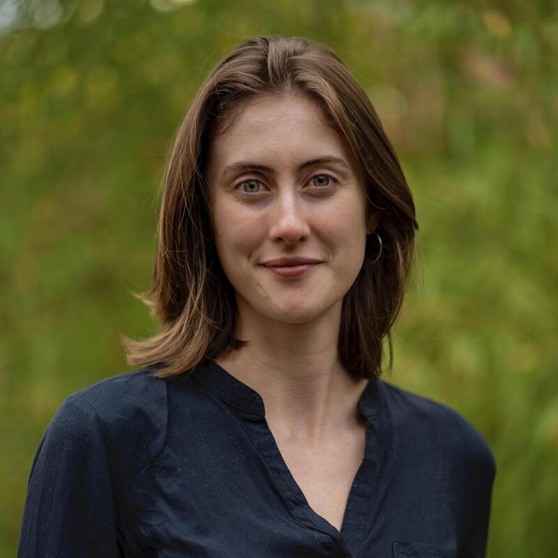 Pale woman with auburn hair in a black shirt standing in front of a green, leafy background. 
