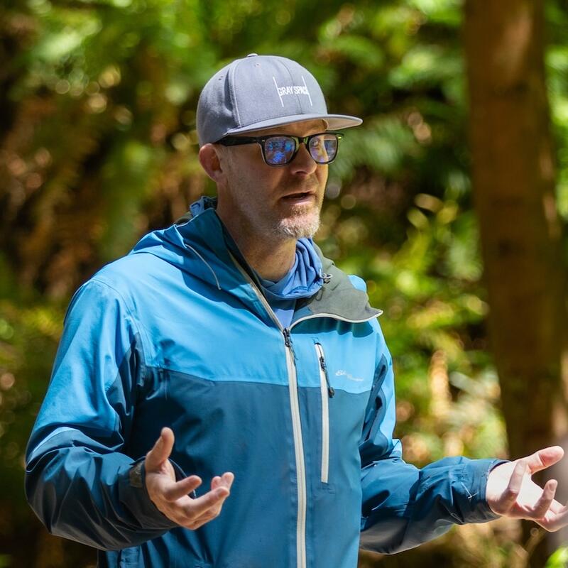 Michael Boonstra speaking outside in a forest wearing a hat and blue jacket during a Creative Field Work course.