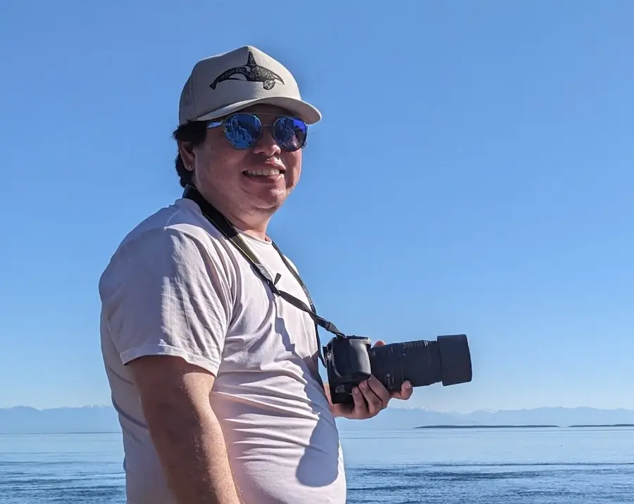 person wearing a white shirt and hat smiling at the camera with a camera hanging around their neck