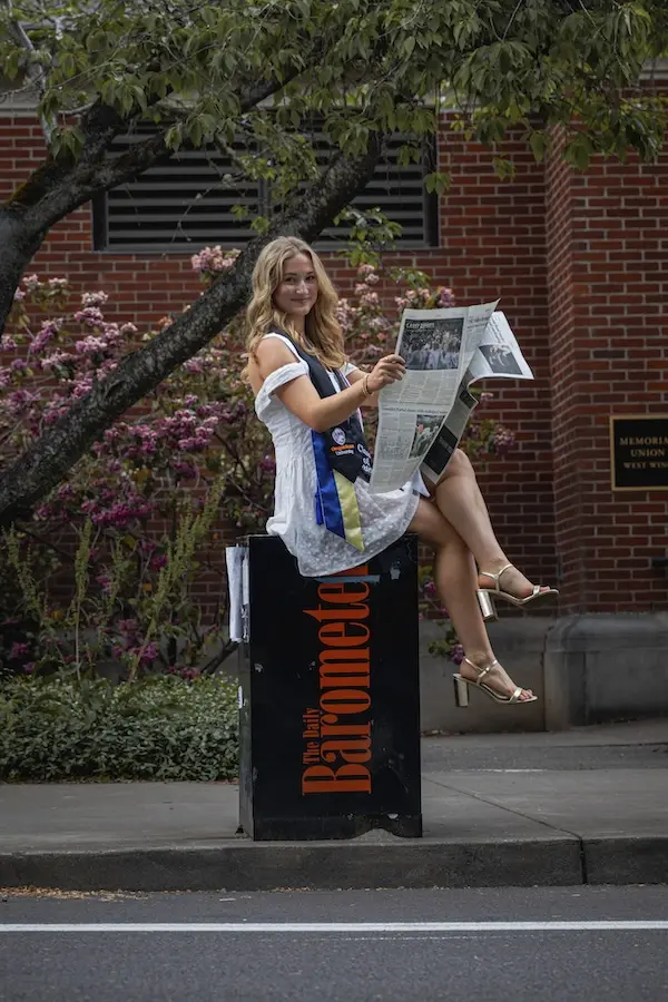 woman in graduation apparel siting on a newspaper stand reading a newspaper