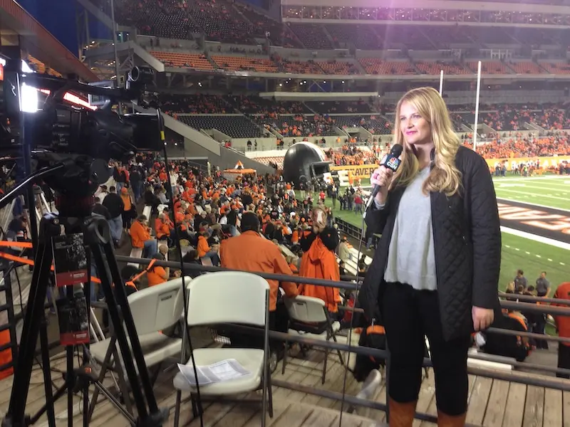 woman speaking into a microphone in front of a television camera at a football game