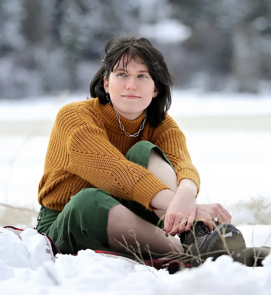 person wearing an orange shirt, green pants, and boots sitting in the snow and looking off into the distance