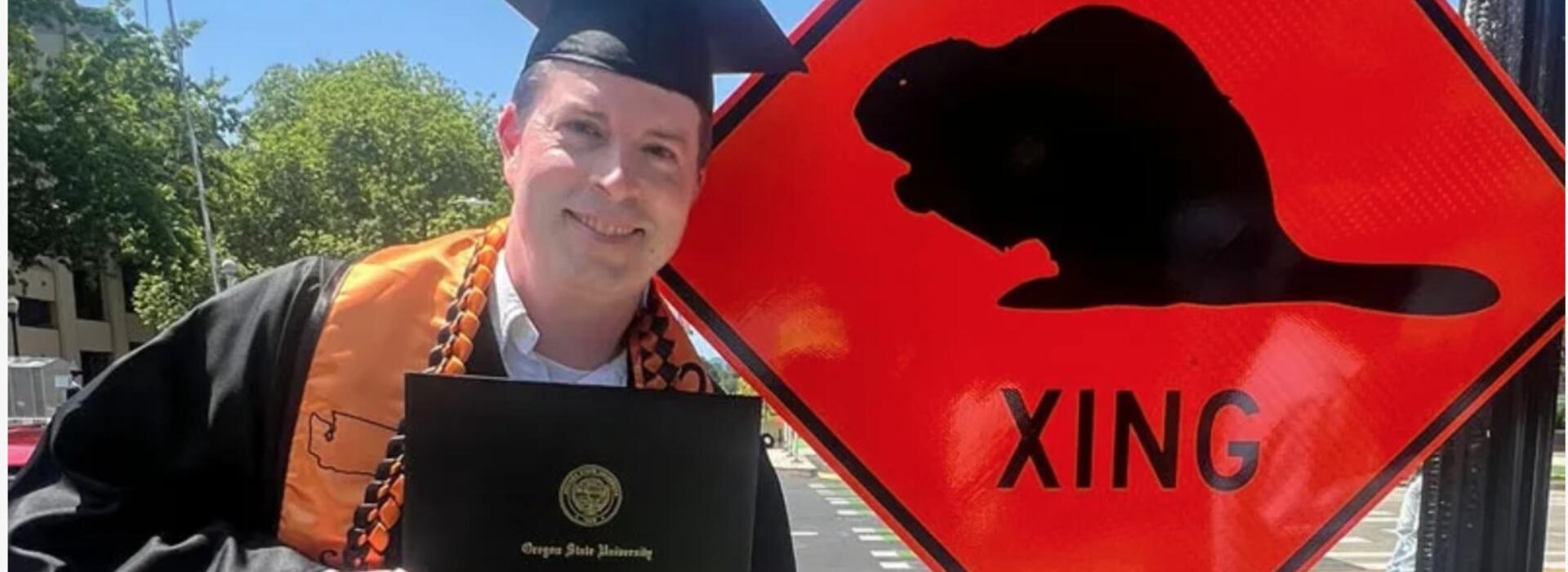 man in graduation cap and gown poses next to beaver crossing sign