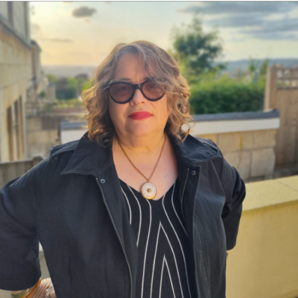 A woman in a striped blouse with wavy, shoulder-length hair and sunglasses stands on a balcony with the sunset behind her